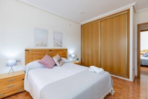 Quarto de hotel com cabeceira de madeira, cama com roupa de cama branca, mesa de cabeceira com abajur, guarda-roupa de madeira e pequena mesinha de cabeceira com uma planta.