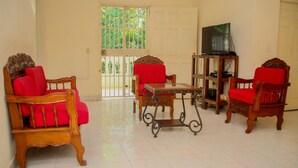 A living room with wooden furniture, a television on a stand, and a view through a glass door to a balcony. - Vacation rental house (Arrondissement de Jacmel)