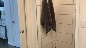 Ein Badezimmer mit weiß gefliesten Wänden, einem Duschbereich mit sechseckischem Bodenmuster und zwei Handtüchern, die an Haken hängen.