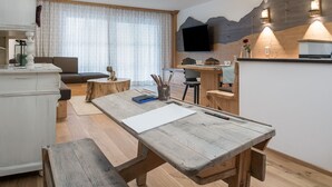 Een rustieke houten eettafel met een witte placemat, een bank en een kleine bijzettafel met een vaas en potloden. Een televisie is bevestigd aan een houten muur, en een bankstel is in de achtergrond zichtbaar.