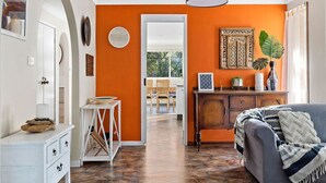 A room with an orange wall, a grey sofa, a wooden cabinet, and a dining area in the background.