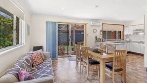 A modern kitchen with white cabinets, a dining area with wooden chairs, and a large window overlooking a garden.