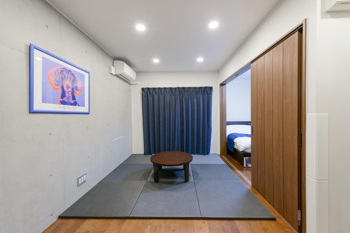 A compact room with a tatami mat floor, a small tatami mat table, a bed with a wooden headboard, a blue curtain, and a framed picture on the wall.