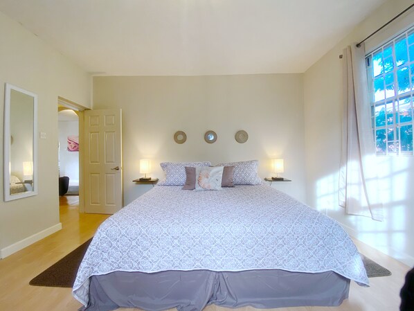 A bedroom with a large bed, two bedside lamps, a mirror, and a window with curtains. - Large 1 Bed + FREE amenities + Close to everything (Bridgetown)