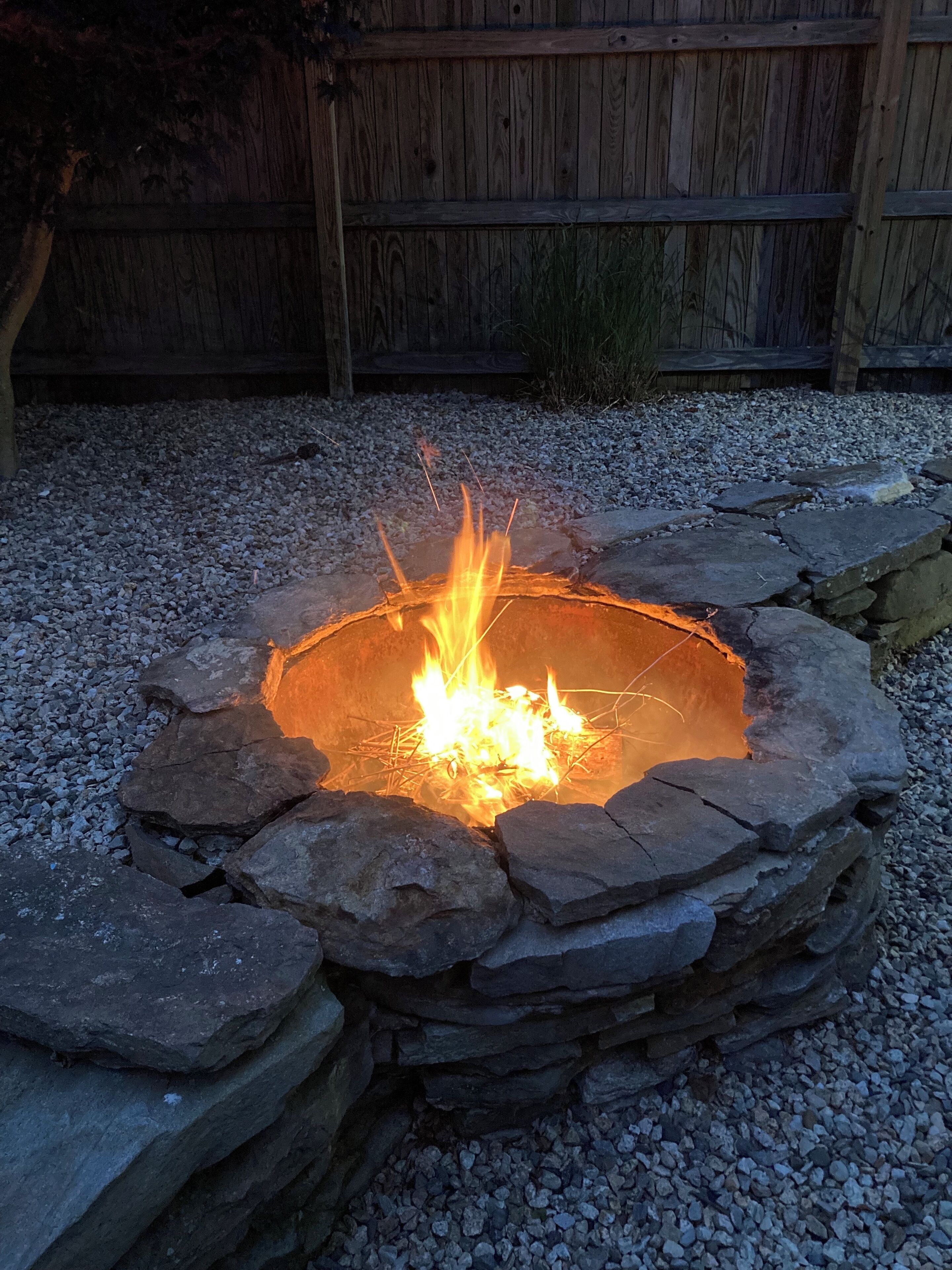 A stone fire pit with a fire burning inside, surrounded by a gravel area and a wooden fence in the background.