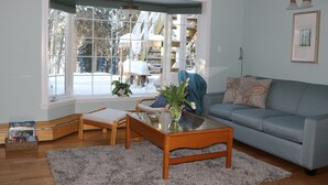Un salon avec une table basse en verre, un canapé, un fauteuil et une grande fenêtre donnant sur une terrasse en bois et des arbres couverts de neige.