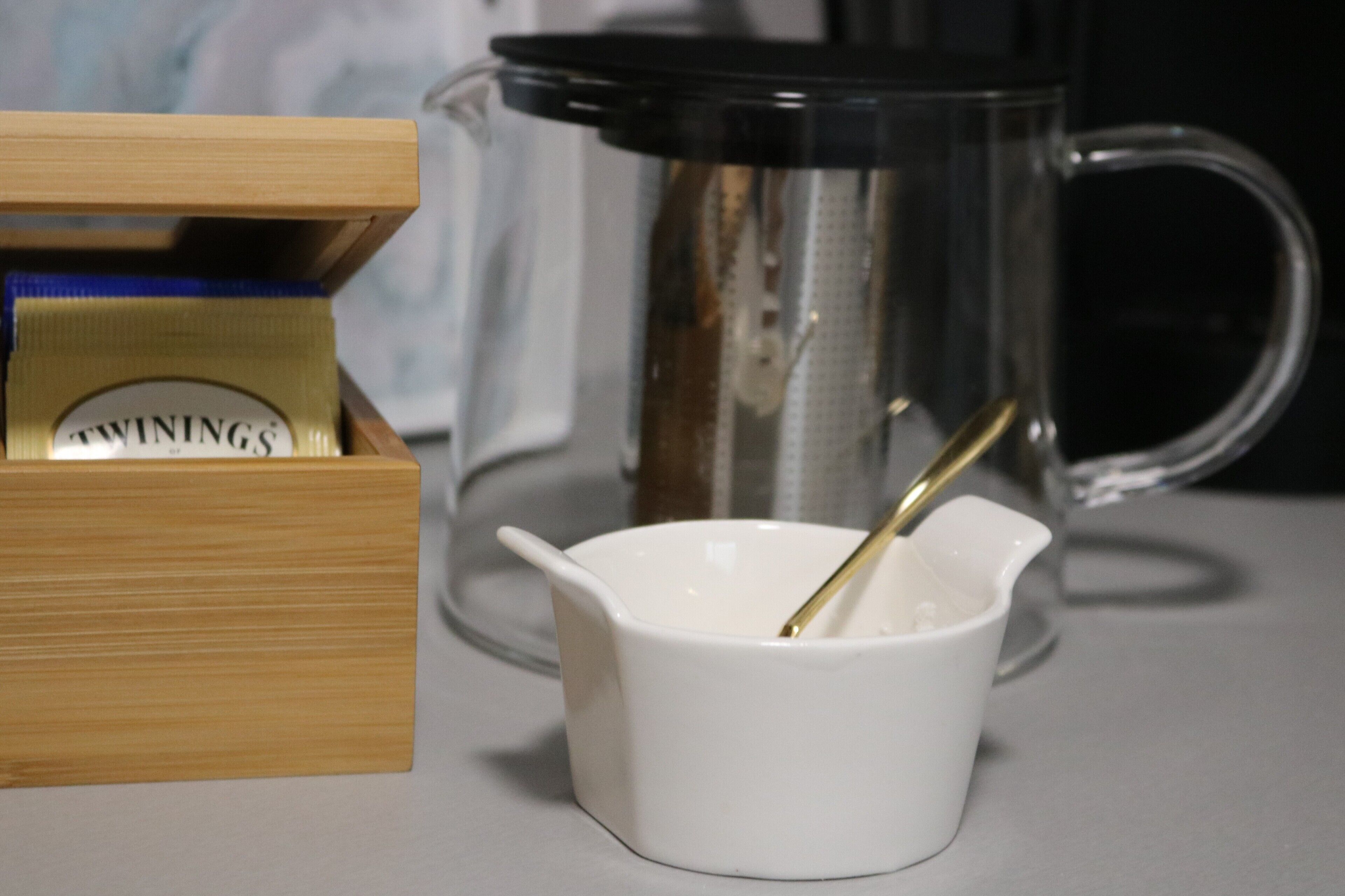 Een witte keramische maatbeker met een gouden lepel, een glazen koffiepot en een houten doos met de merknaam 'TWININGS' op een tafel.