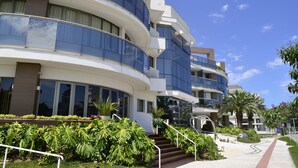 A modern multi-story hotel with curved balconies, large windows, and a landscaped garden area with palm trees and shrubs. - Apartamento de 2 Quartos no Luxuoso Condomínio Jay (Jurerê Internacional)