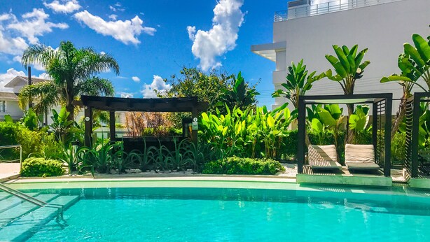 A swimming pool with a pergola and lounge chairs, surrounded by tropical plants and a clear blue sky. - Apartamento de 2 Quartos no Luxuoso Condomínio Jay (Jurerê Internacional)