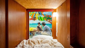 Una habitación con una cama, paredes de madera, un cuadro de una playa y una mesa con un gato y una persona.
