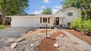 Une maison de plain-pied avec un garage, un porche à l’avant doté d’un banc, et un jardin aménagé avec du paillis et des rochers.