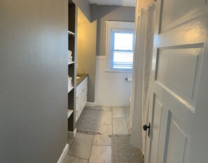 A narrow hallway with a door leading to a room with a window. There are shelves with towels and a rug on the floor.