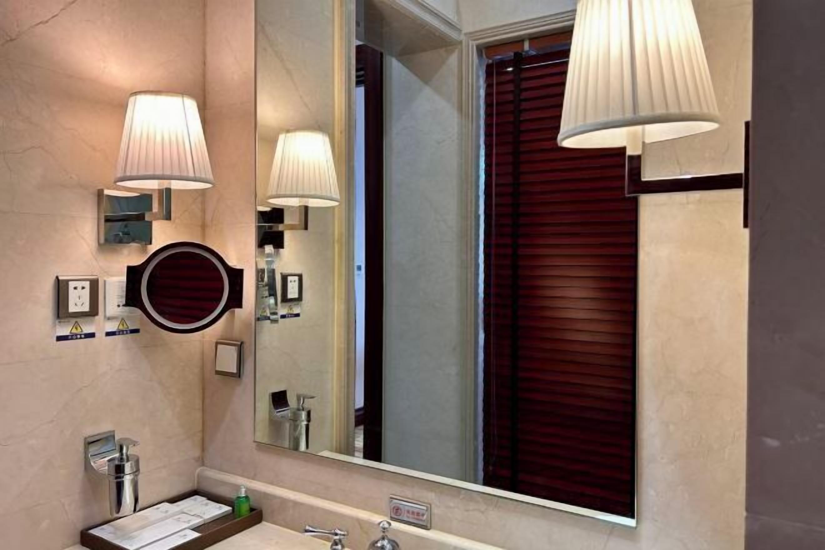 A bathroom with a large mirror, wall-mounted lights, a round mirror, and a window with blinds.