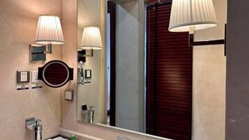 A bathroom with a large mirror, wall-mounted lights, a round mirror, and a window with blinds.