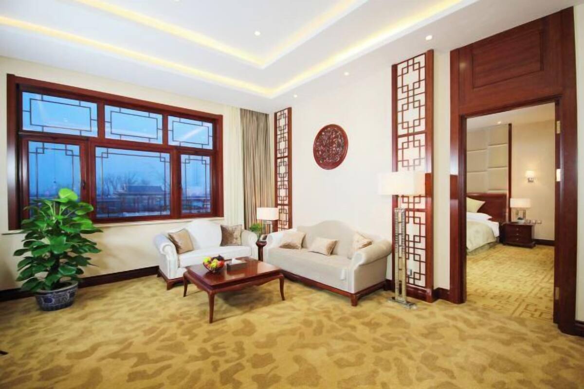 A hotel room with a sofa, a coffee table, a potted plant, a decorative screen, and a bed with a nightstand.
