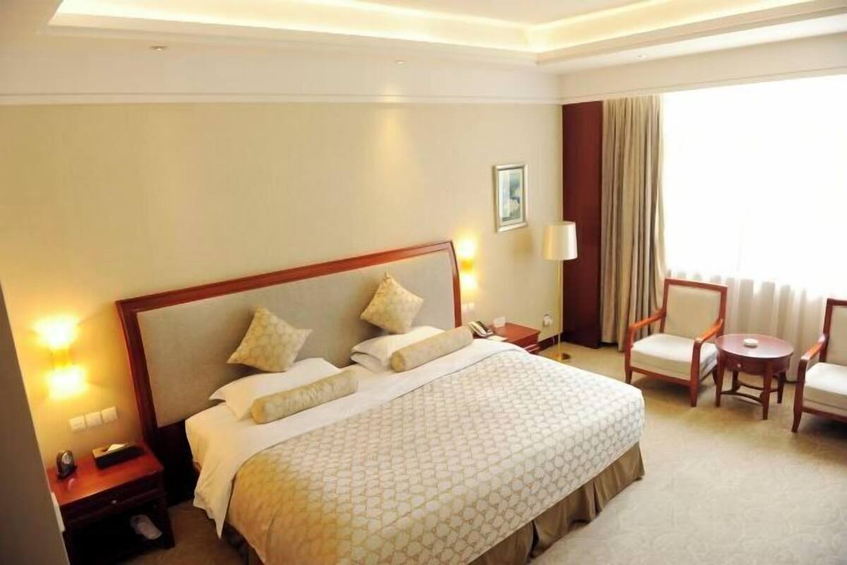 A hotel room with a large bed, bedside tables, a chair, a small table, and a window with curtains.