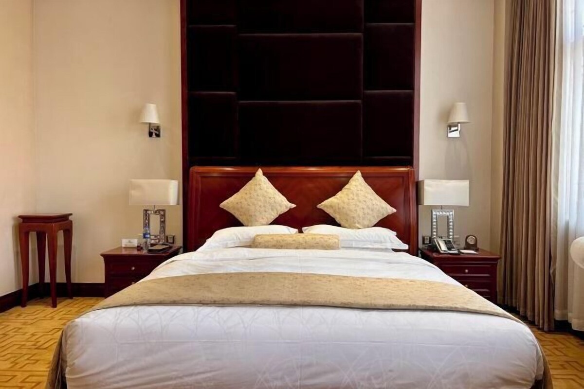 Une chambre d’hôtel avec un grand lit, deux tables de chevet, une tête de lit et une fenêtre avec des rideaux.