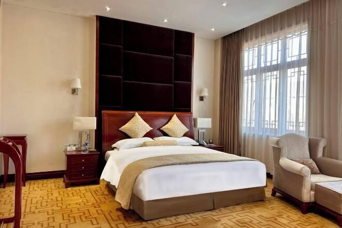 A hotel room with a large bed, a wooden headboard, a nightstand, a desk lamp, a chair, and a window with curtains.