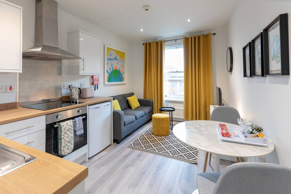 A modern kitchen with a stove, oven, and sink. There is a gray sofa with yellow pillows, a round table with chairs, and a window with yellow curtains.