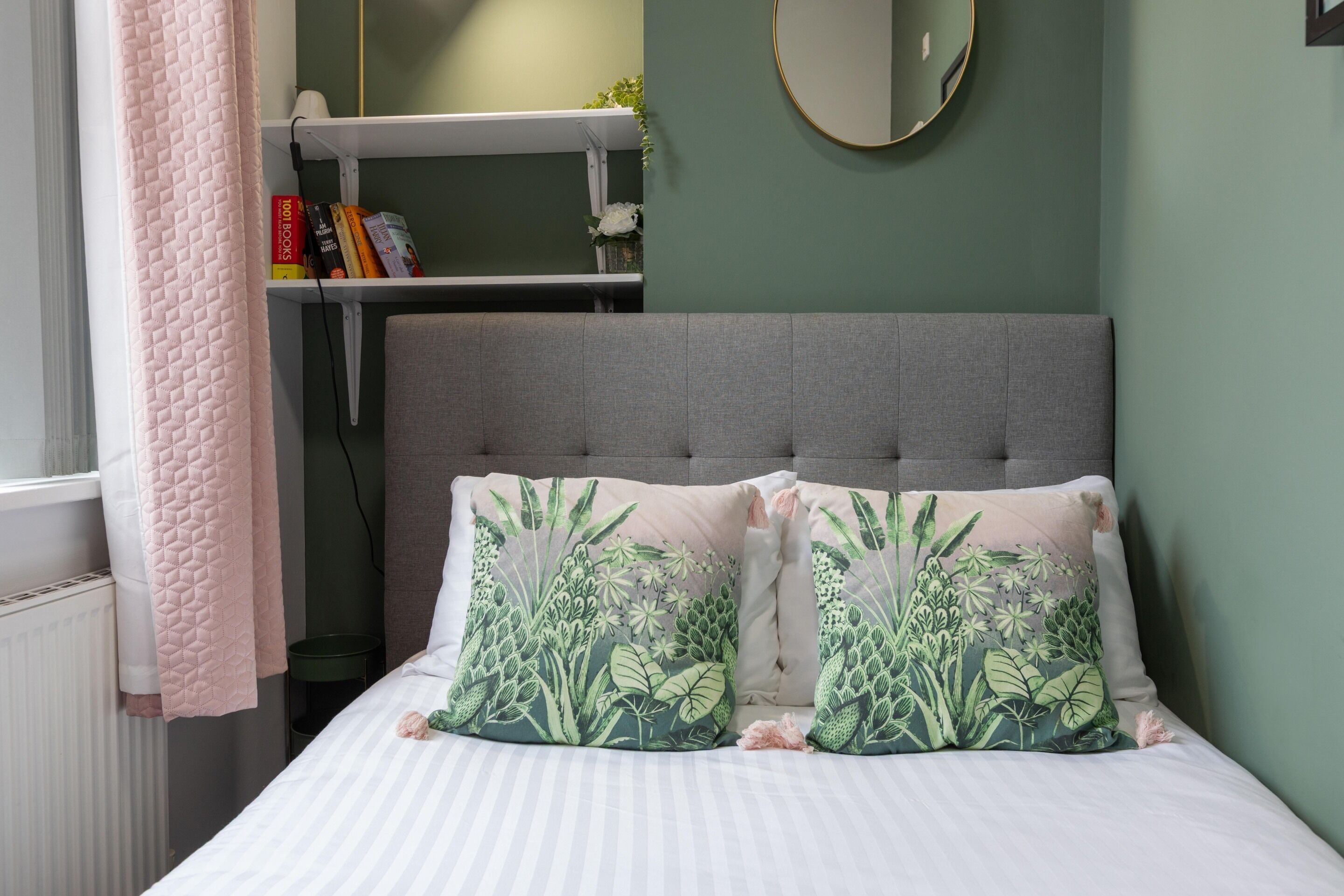A bedroom with a grey upholstered headboard, two floral-patterned pillows, a white-striped bedspread, and a pink quilted blanket.