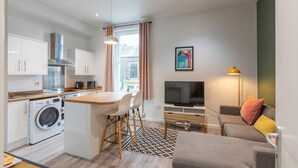 Un appartement moderne comprenant une cuisine, un coin repas et un salon. Il est équipé d’un lave-linge, d’une table à manger avec des chaises, d’un canapé, d’une télévision et d’une lampe.