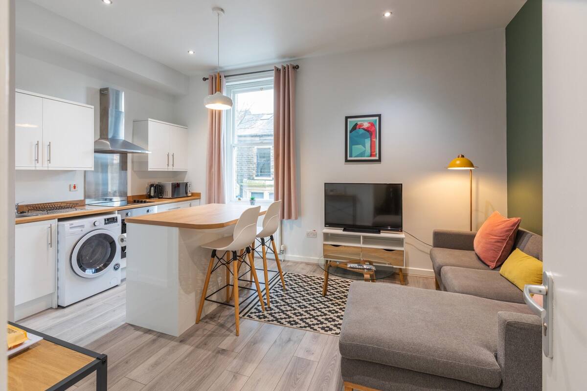 A modern apartment with a kitchen, dining area, and living room. It includes a washing machine, a dining table with chairs, a sofa, a TV, and a lamp.