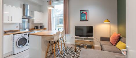 A modern apartment with a kitchen, dining area, and living room. It includes a washing machine, a dining table with chairs, a sofa, a TV, and a lamp.