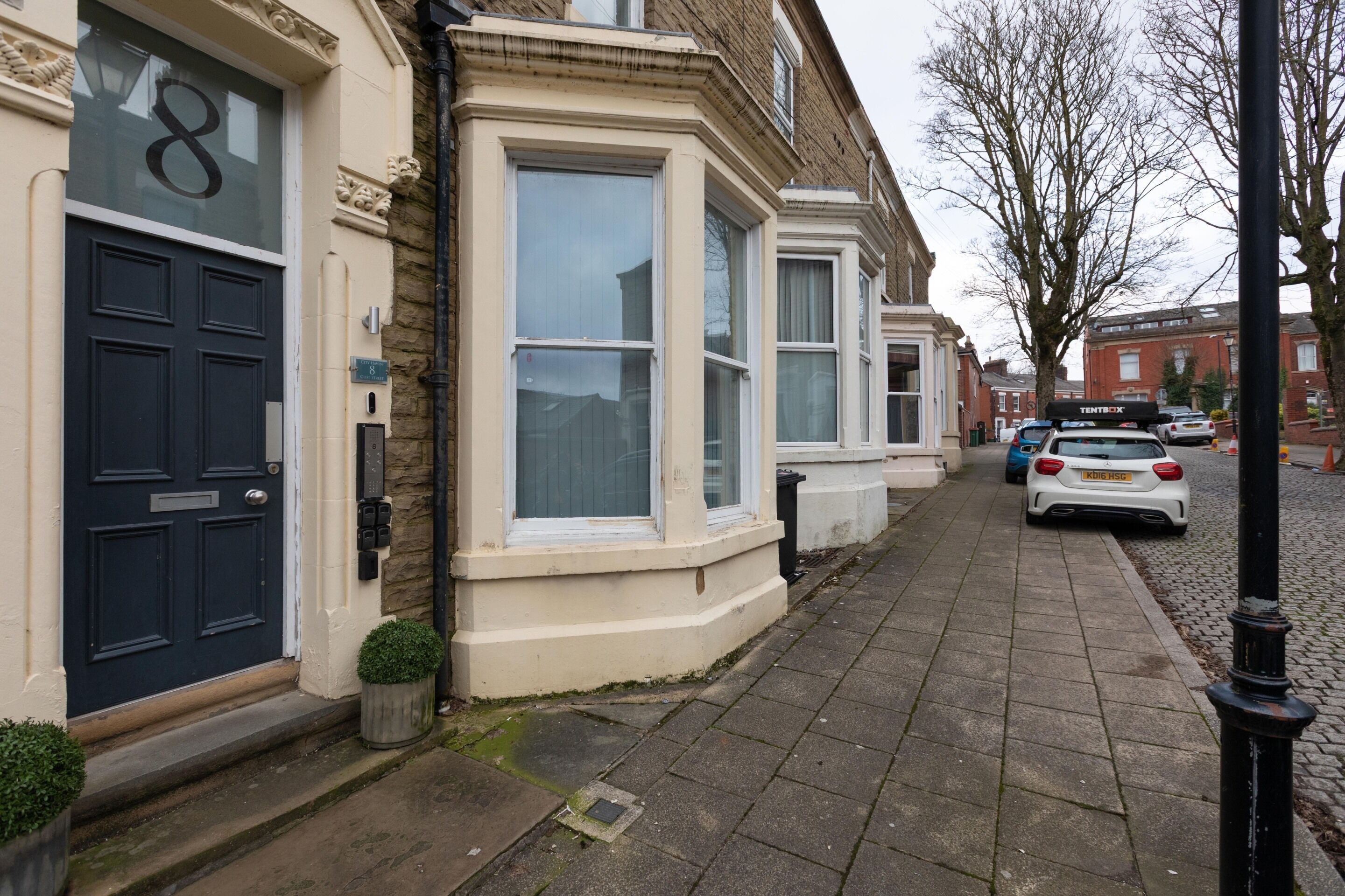 A row of terraced houses with bay windows, a cobblestone street, and parked cars.