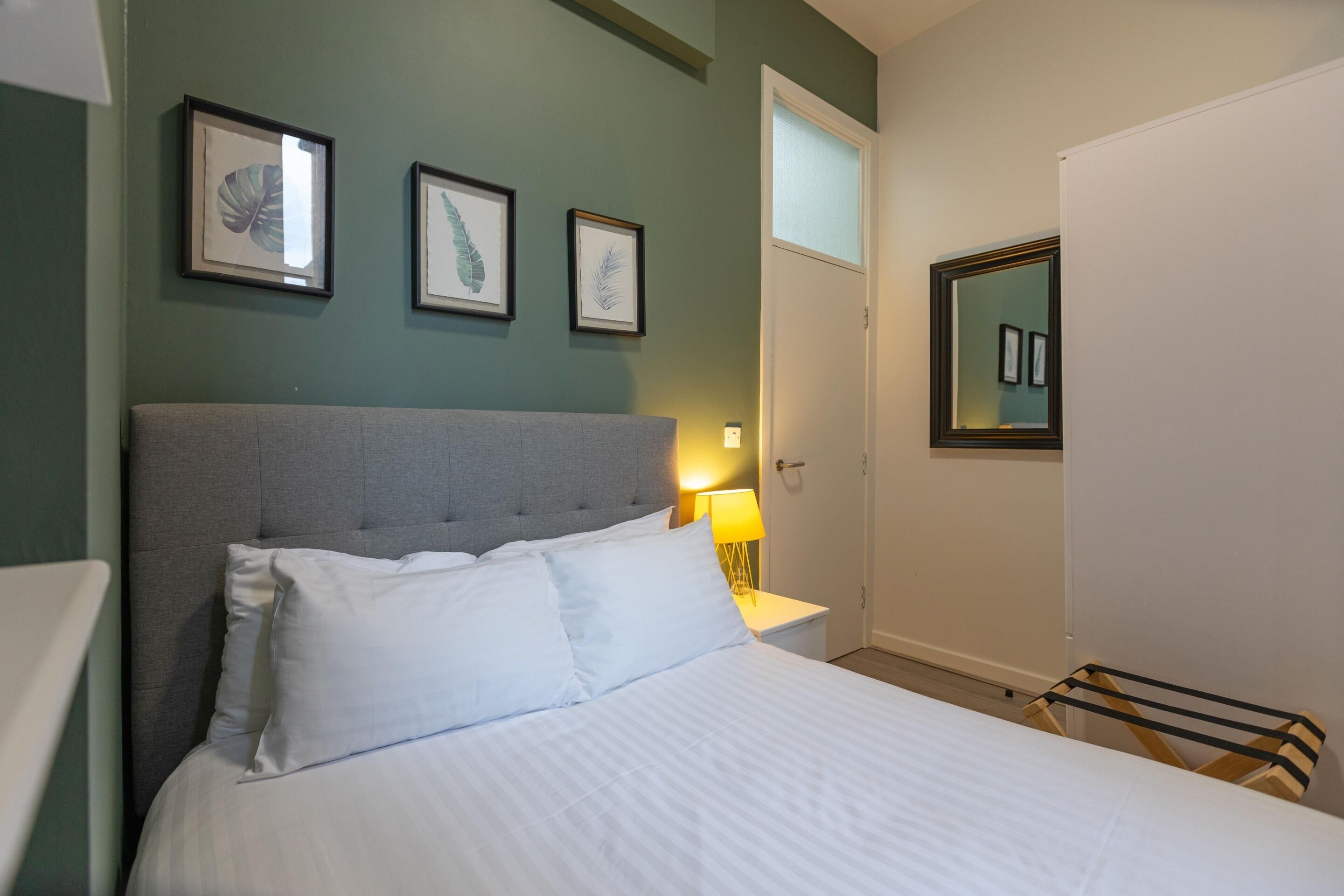 Una cama bien hecha con ropa blanca en una habitación con paredes verdes, cabecera gris y obras de arte enmarcadas en la pared.