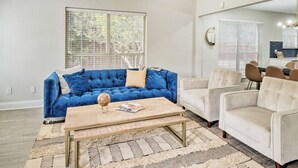 Un salon comprenant un canapé bleu, un canapé beige, une table basse et un coin repas avec des chaises.