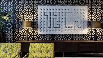 Un vestíbulo con una pared de diseño geométrico, dos sillones con estampado floral y una gran pantalla numérica iluminada.