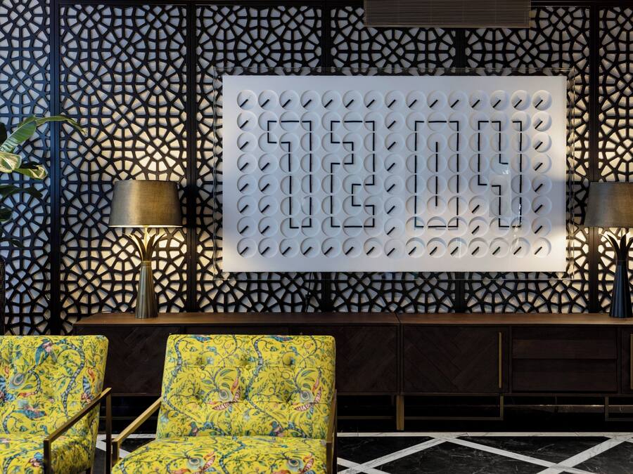Un vestíbulo con una pared de diseño geométrico, dos sillones con estampado floral y una gran pantalla numérica iluminada.