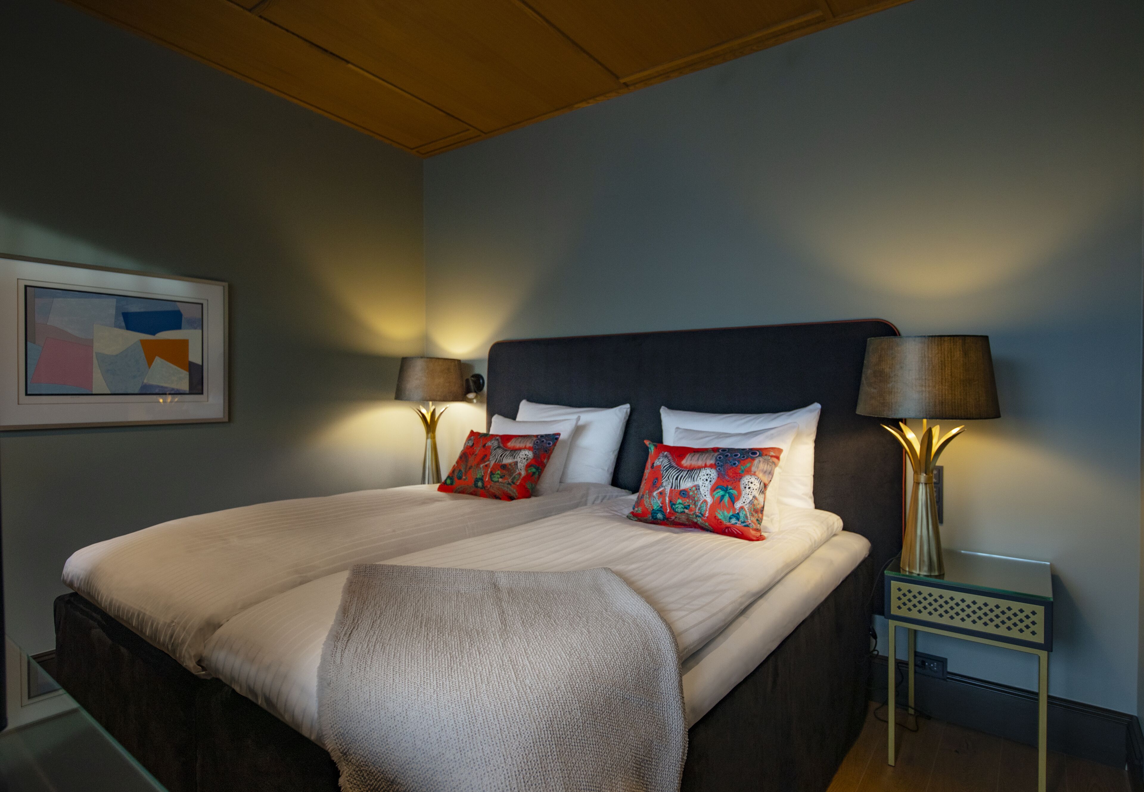 A neatly made bed with two pillows, a bedside table with a lamp, and a framed abstract painting on the wall.