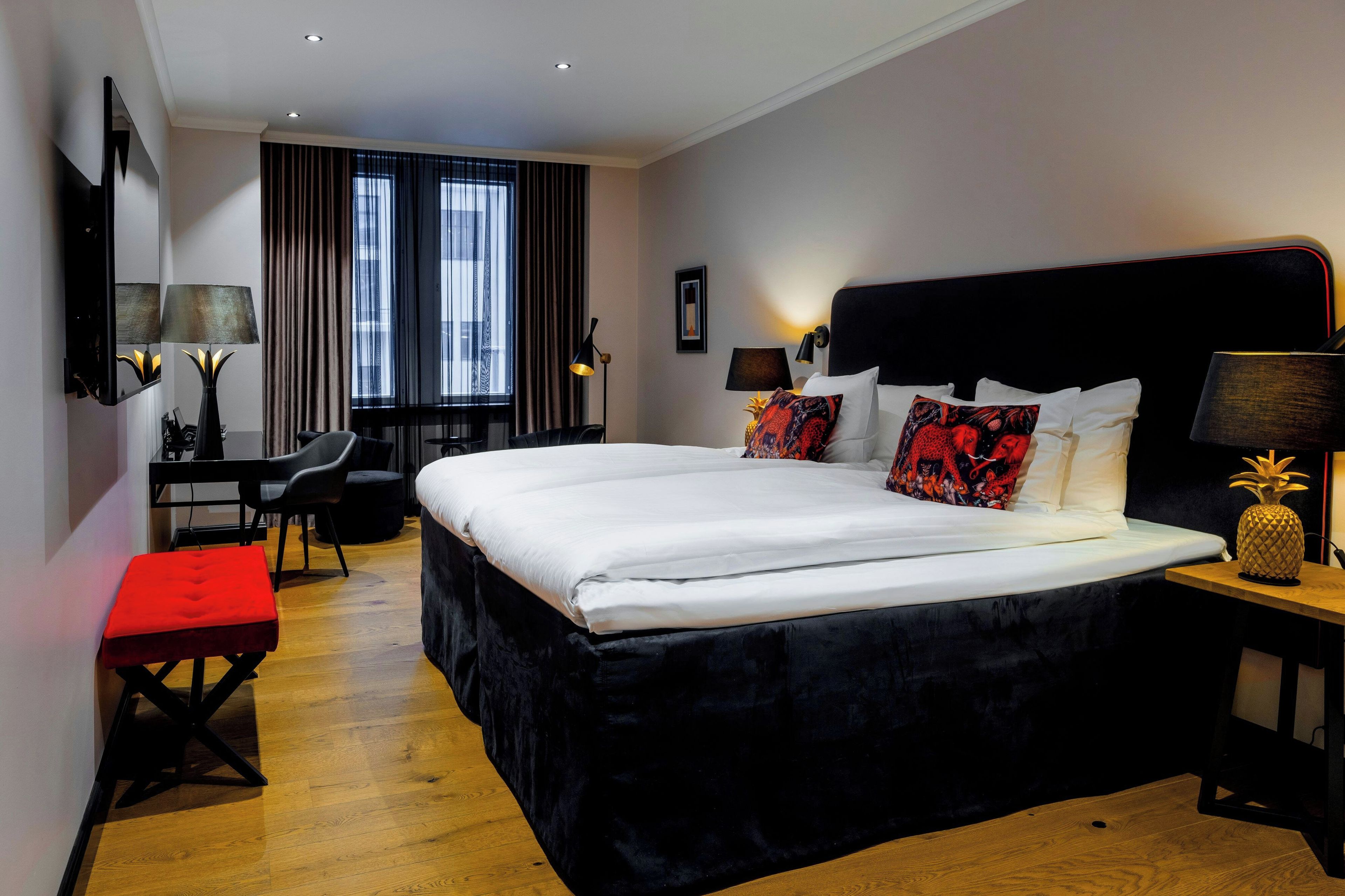 A hotel room with a large bed, a desk, a chair, a red ottoman, and a nightstand with a lamp.