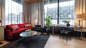 Une chambre d’hôtel moderne avec un canapé rouge, une table basse ronde, une grande plante en pot et un bureau avec une lampe.