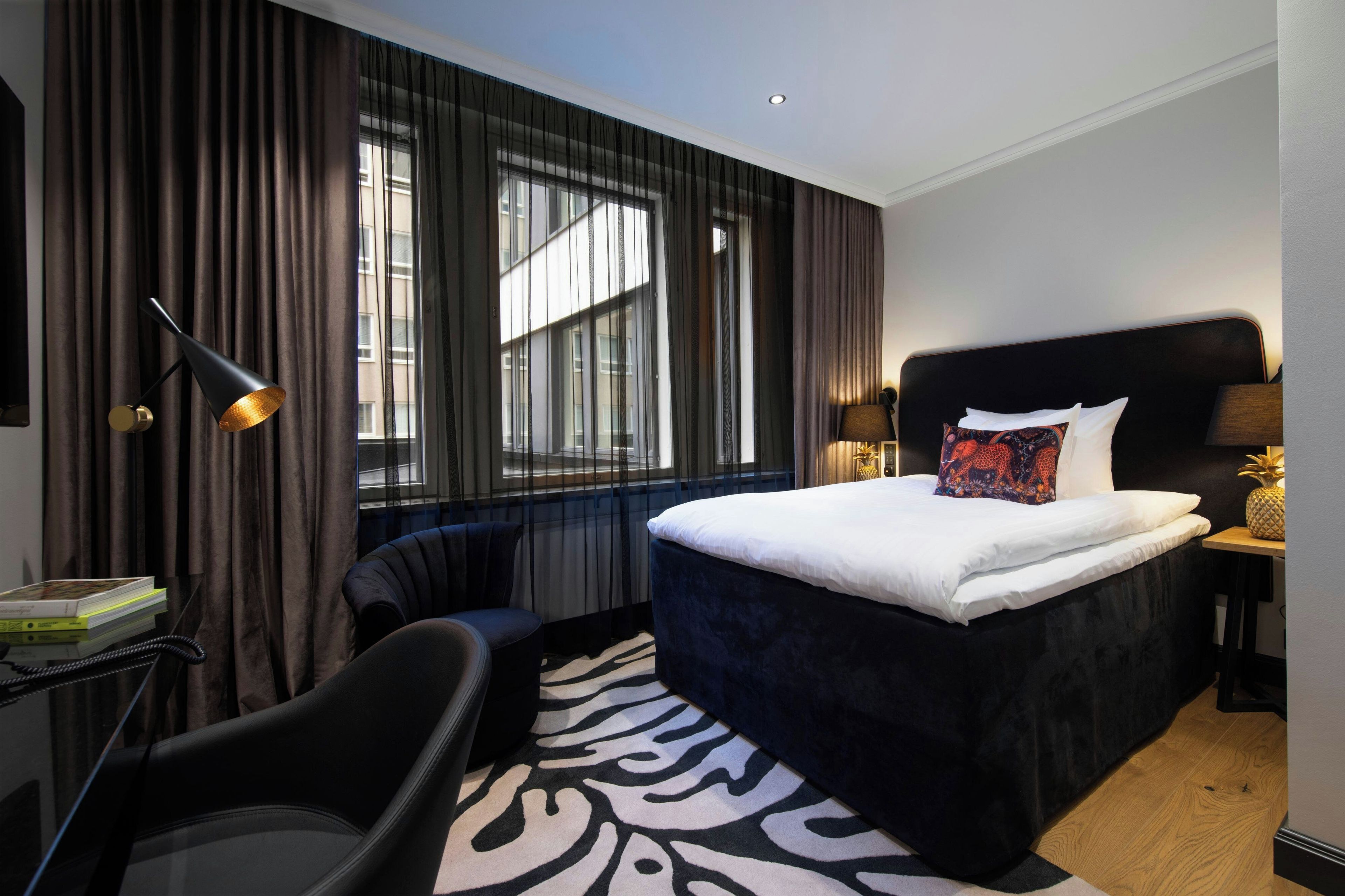 A hotel room with a large bed, a desk with a lamp, a chair, and a window with curtains.