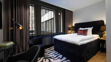 A hotel room with a large bed, a desk with a lamp, a chair, and a window with curtains.
