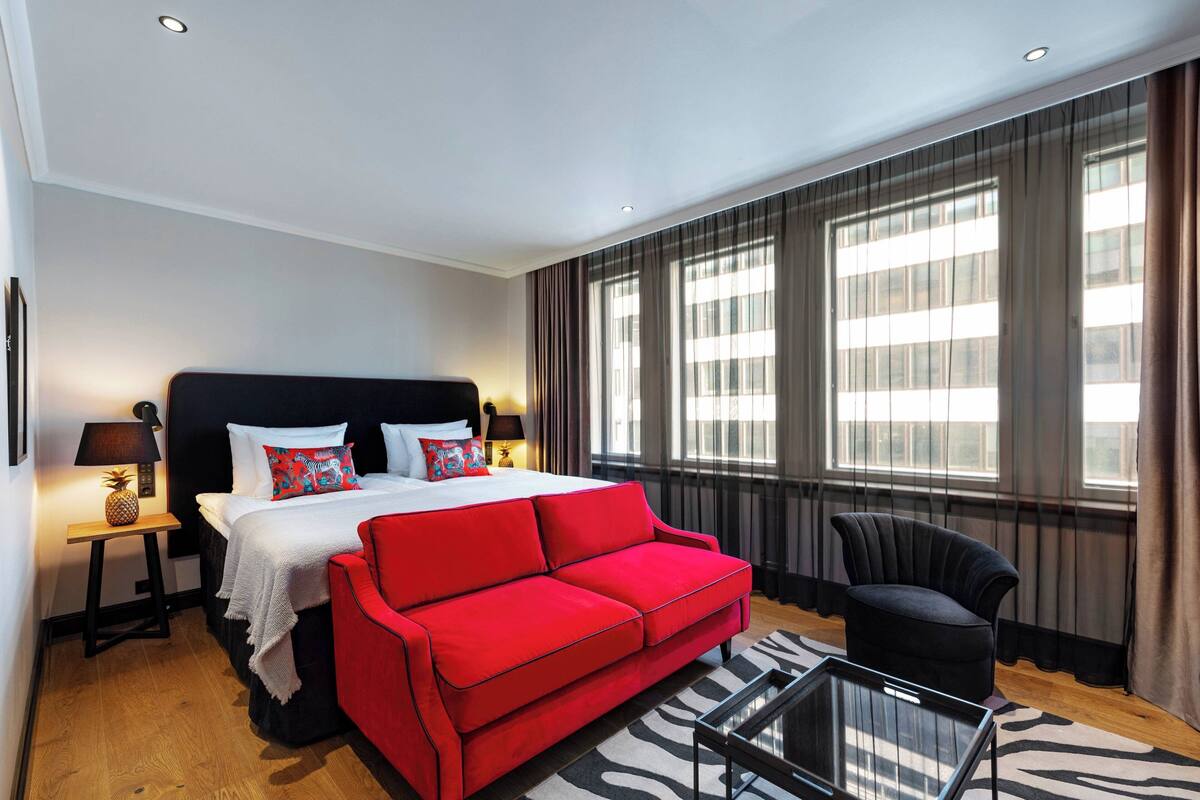 Camera d'albergo con un letto grande, un divano rosso, una poltrona nera, un tavolino da caffè in vetro e un tappeto a motivi zebrati.