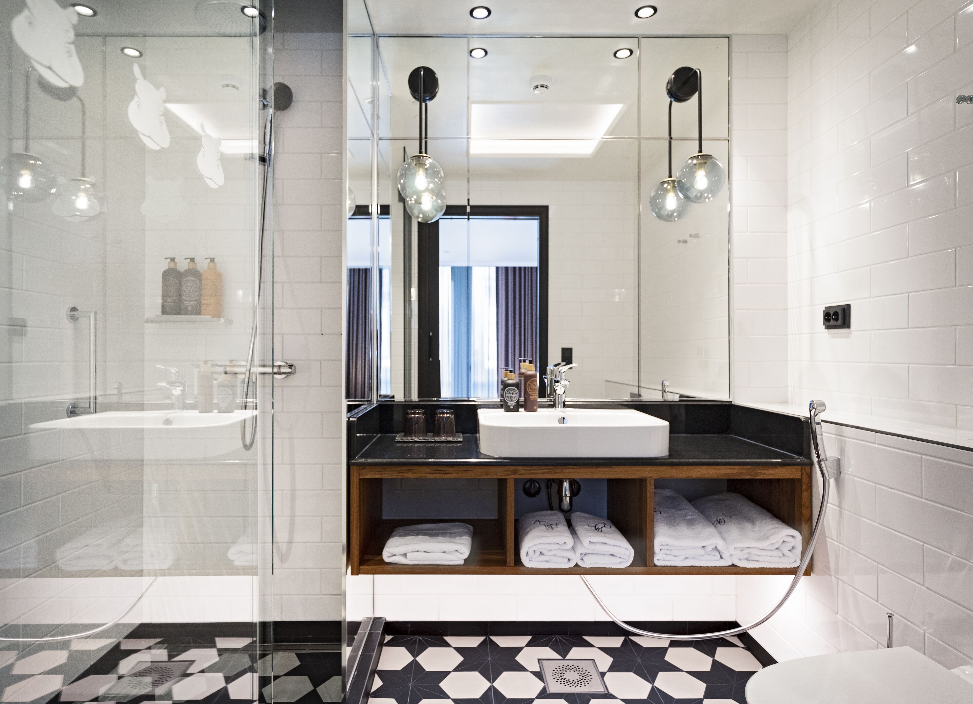 Une salle de bain moderne avec une douche à l’italienne en verre, un grand miroir, un lavabo avec un meuble-vasque en bois et un carrelage décoratif.