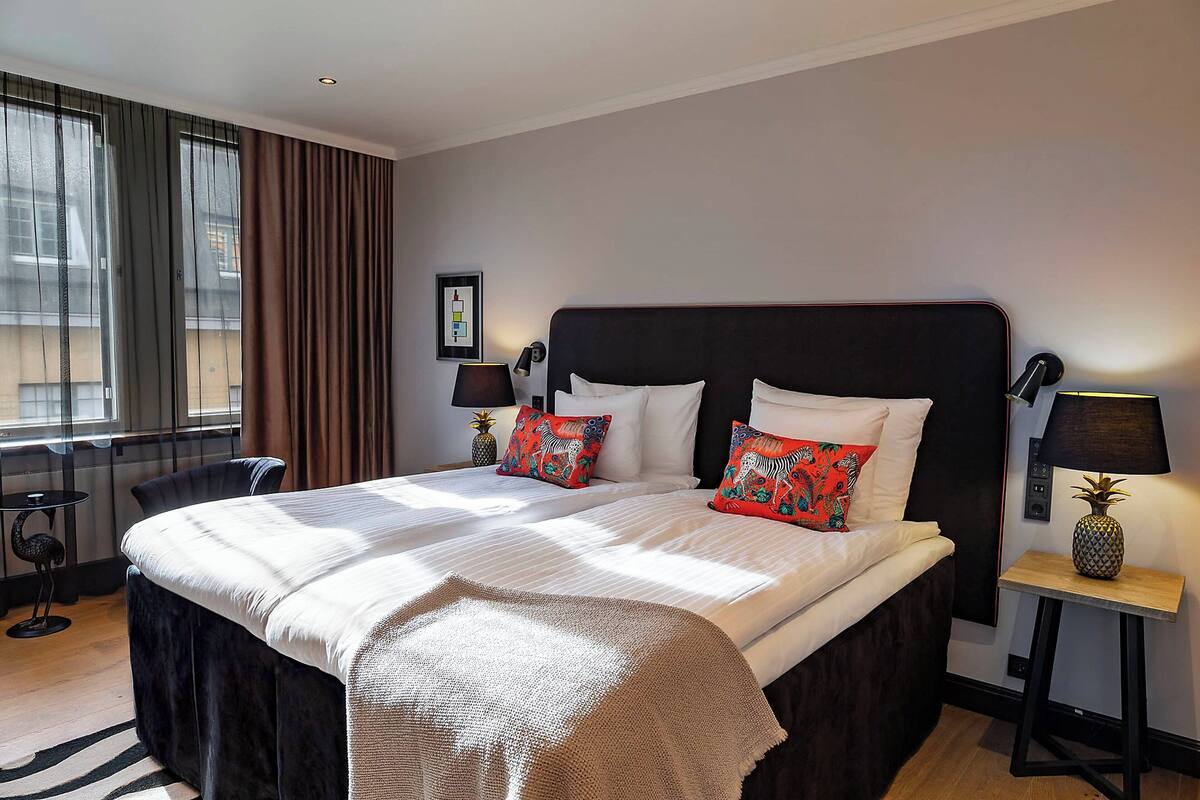Una camera d'hotel con un letto grande, due comodini, un tavolino, una sedia e una finestra con tende.