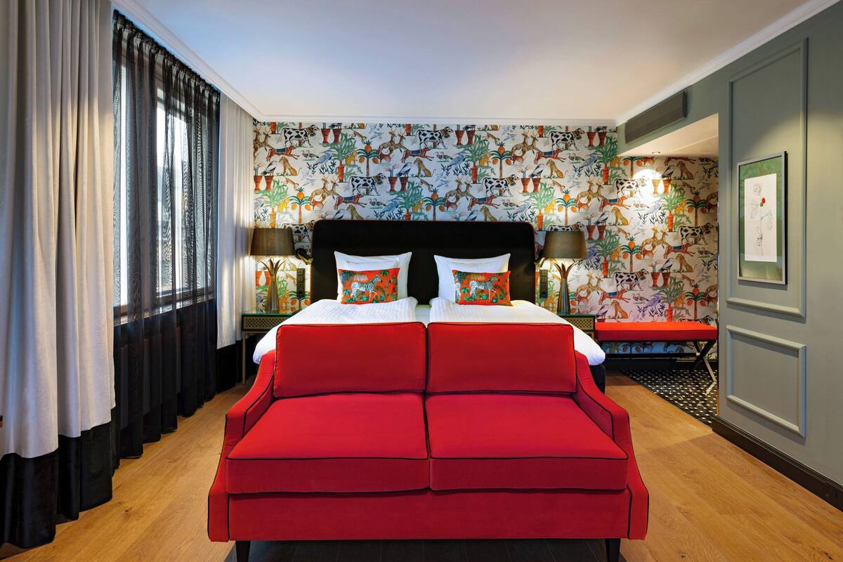 Una camera d'albergo con un letto grande, un divano rosso, carta da parati a motivi e una panca.