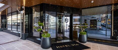 L’entrée d’un hôtel, avec un paillasson noir sur lequel on peut lire « HÔTEL U14 », des portes en verre et des plantes en pot.