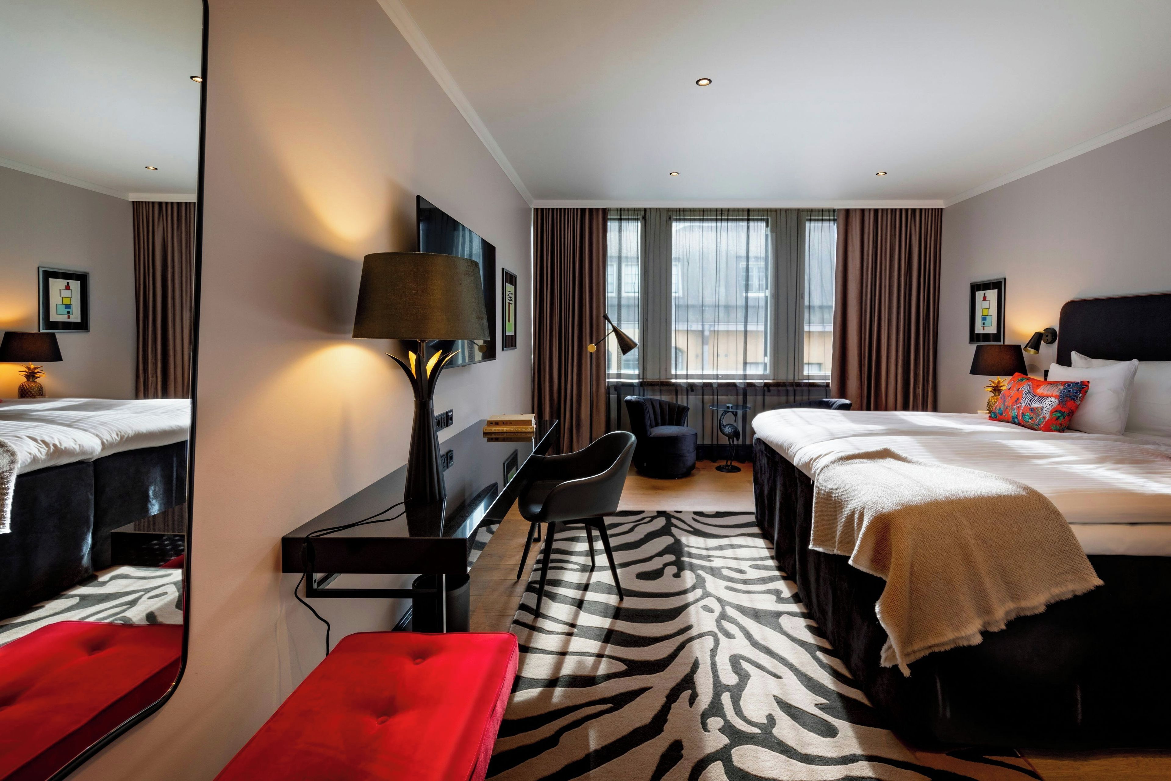 Une chambre d’hôtel avec un lit, un bureau, une chaise, un tapis à motifs zébrés et une fenêtre avec des rideaux.