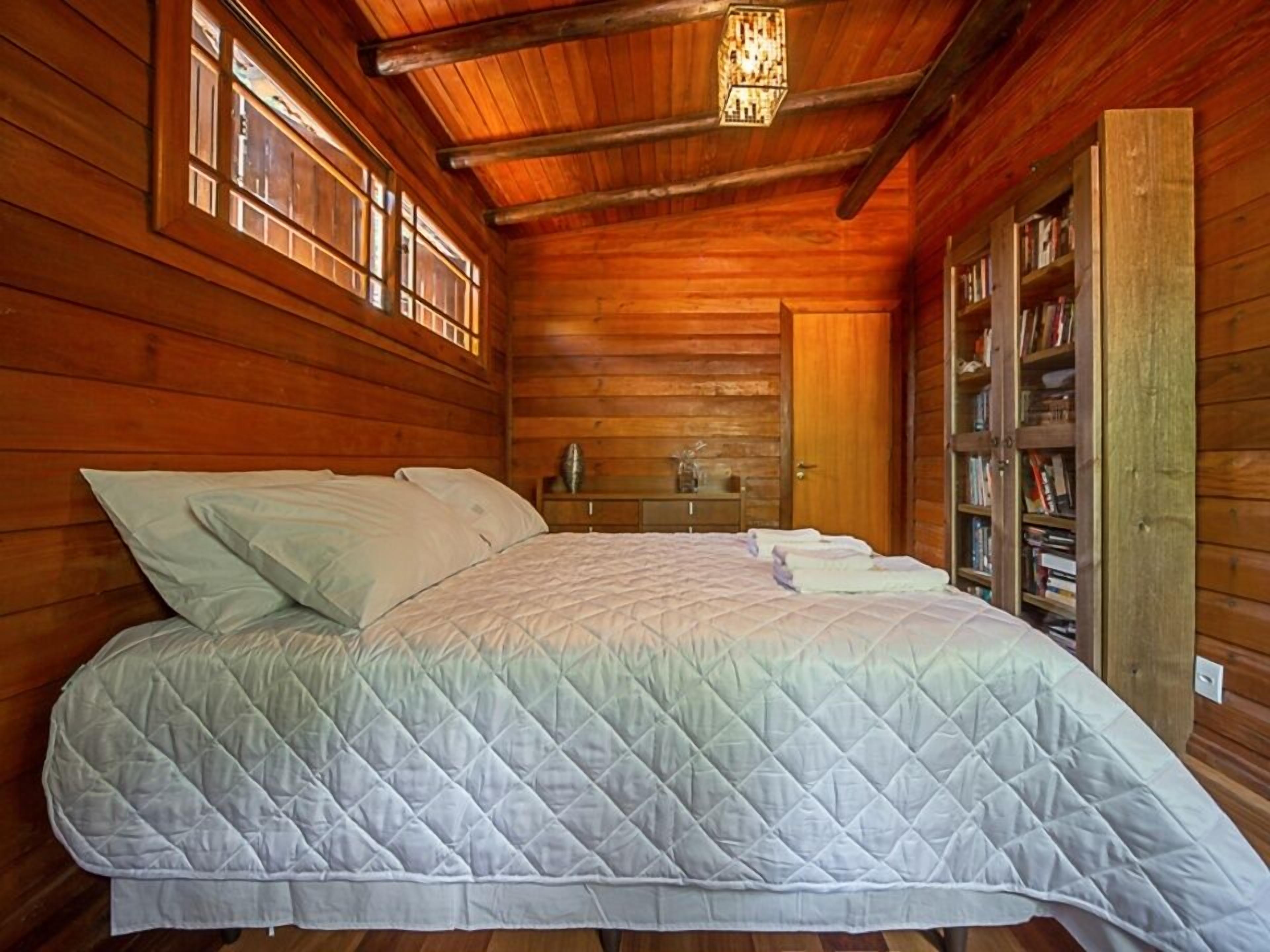 A wooden cabin room with a bed, a nightstand, a bookshelf, and a window.
