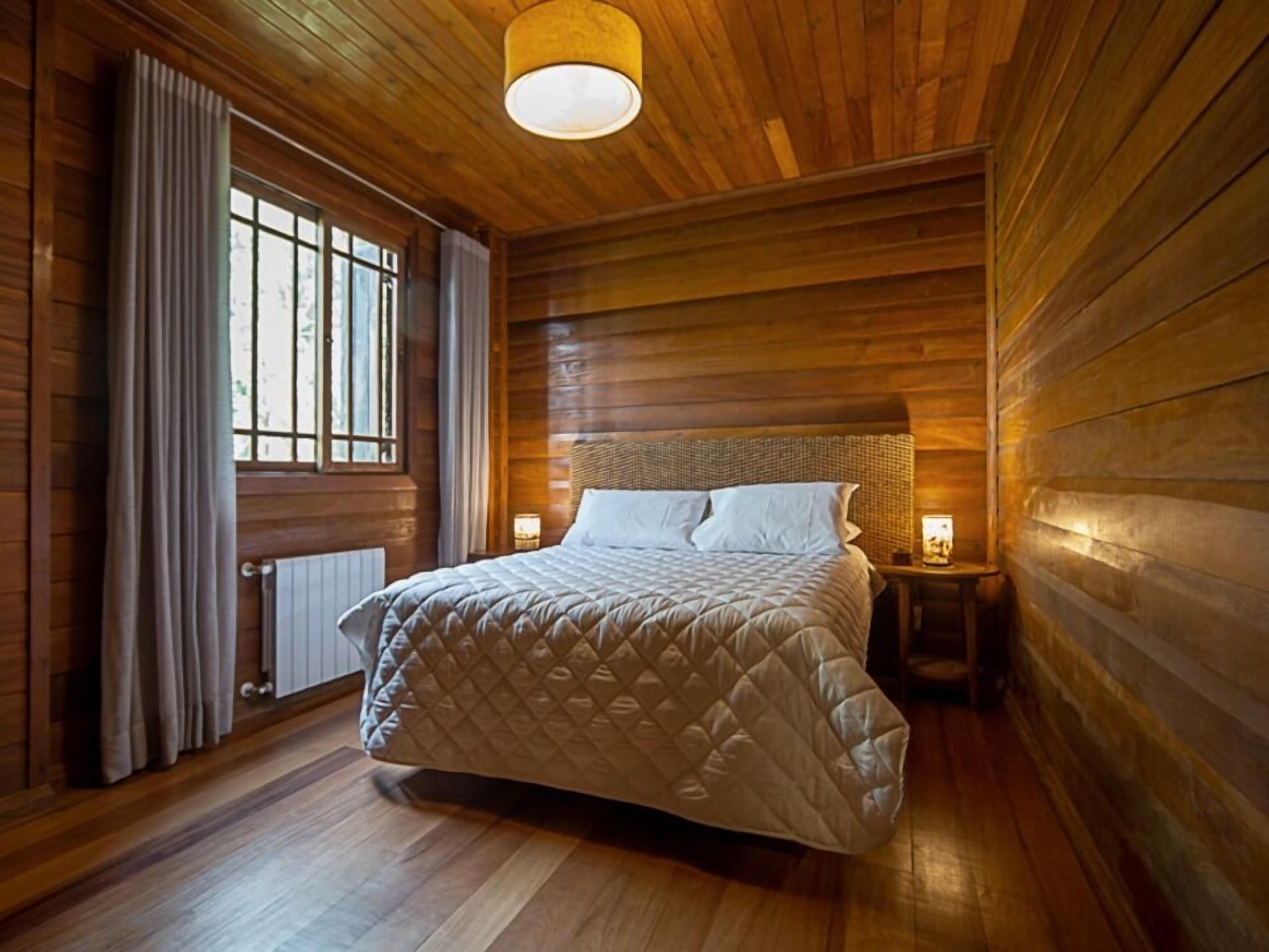 A wooden-paneled bedroom with a bed, a window with curtains, and a nightstand with a lamp.