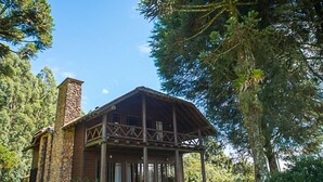 A rustic cabin with a stone chimney, wooden porch, and surrounded by lush greenery. - Aires de Patagônia Pousada (Cambara do Sul)