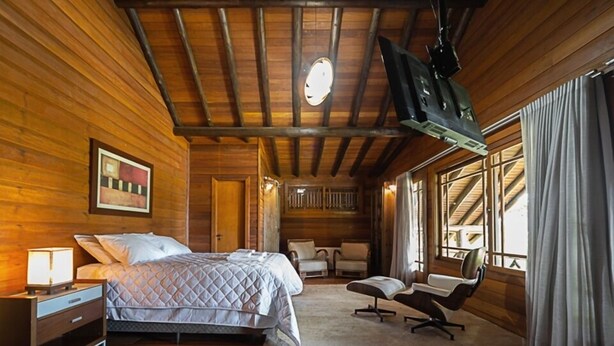 A wooden cabin bedroom with a bed, bedside table, chair, and a large window. - Aires de Patagônia Pousada (Cambara do Sul)