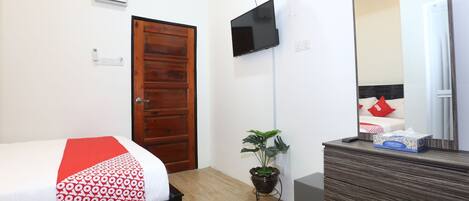 A bedroom with a bed, a wooden door, a TV mounted on the wall, a potted plant, and a dresser with a mirror.