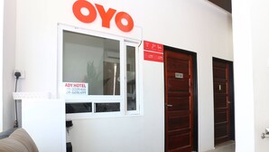 Zona de recepción de hotel con un cartel que indica 'OYO' y 'ADY HOTEL' y números de contacto. Hay una puerta que dice 'SOLO PARA PERSONAL' y un mostrador de recepción con un teléfono y una planta.