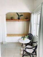 Um quarto pequeno e iluminado, com um banco branco, duas cadeiras pretas, uma mesa redonda e plantas em vasos sobre uma estante.